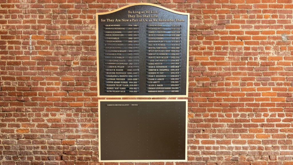Wall-mounted bronze memorial plaque with raised gold lettering and individual name plates on a brick wall