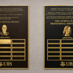 Two custom bronze and gold UBS recognition plaques with engraved portraits, award text, and individual nameplates mounted on an interior wall.