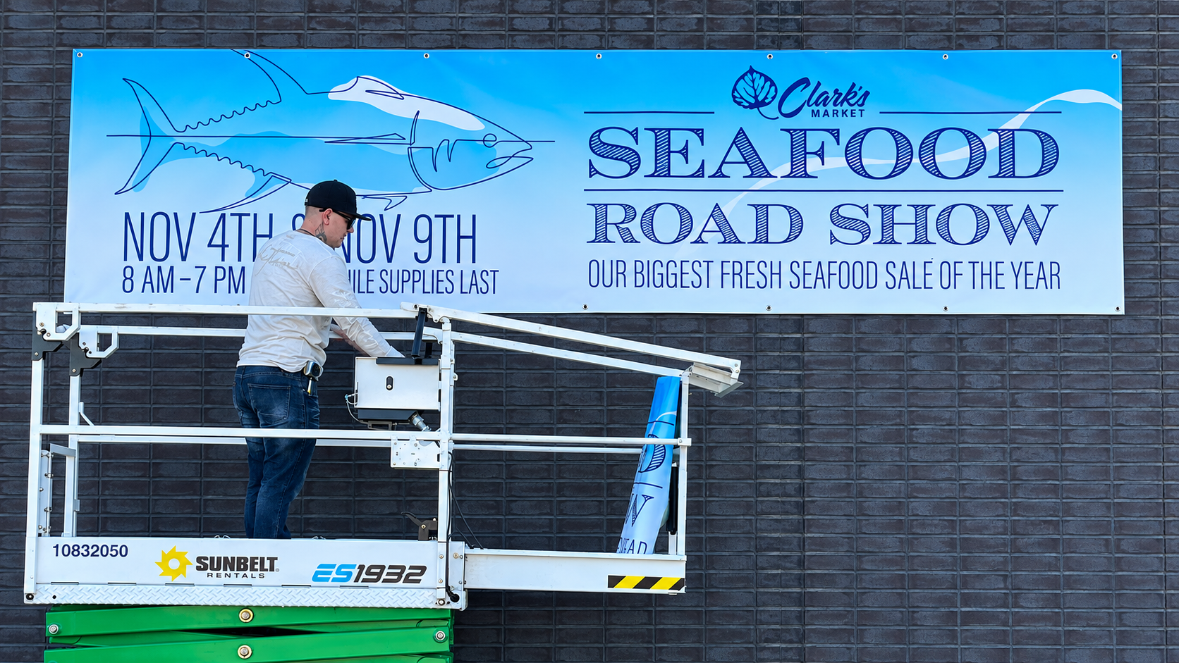 Sign installer on a lift installing a large Clark’s Market Seafood Road Show banner on a brick exterior wall